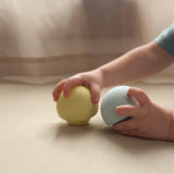 Two hands holding a yellow and a blue tactile silicone montessori ball on a beige surface.